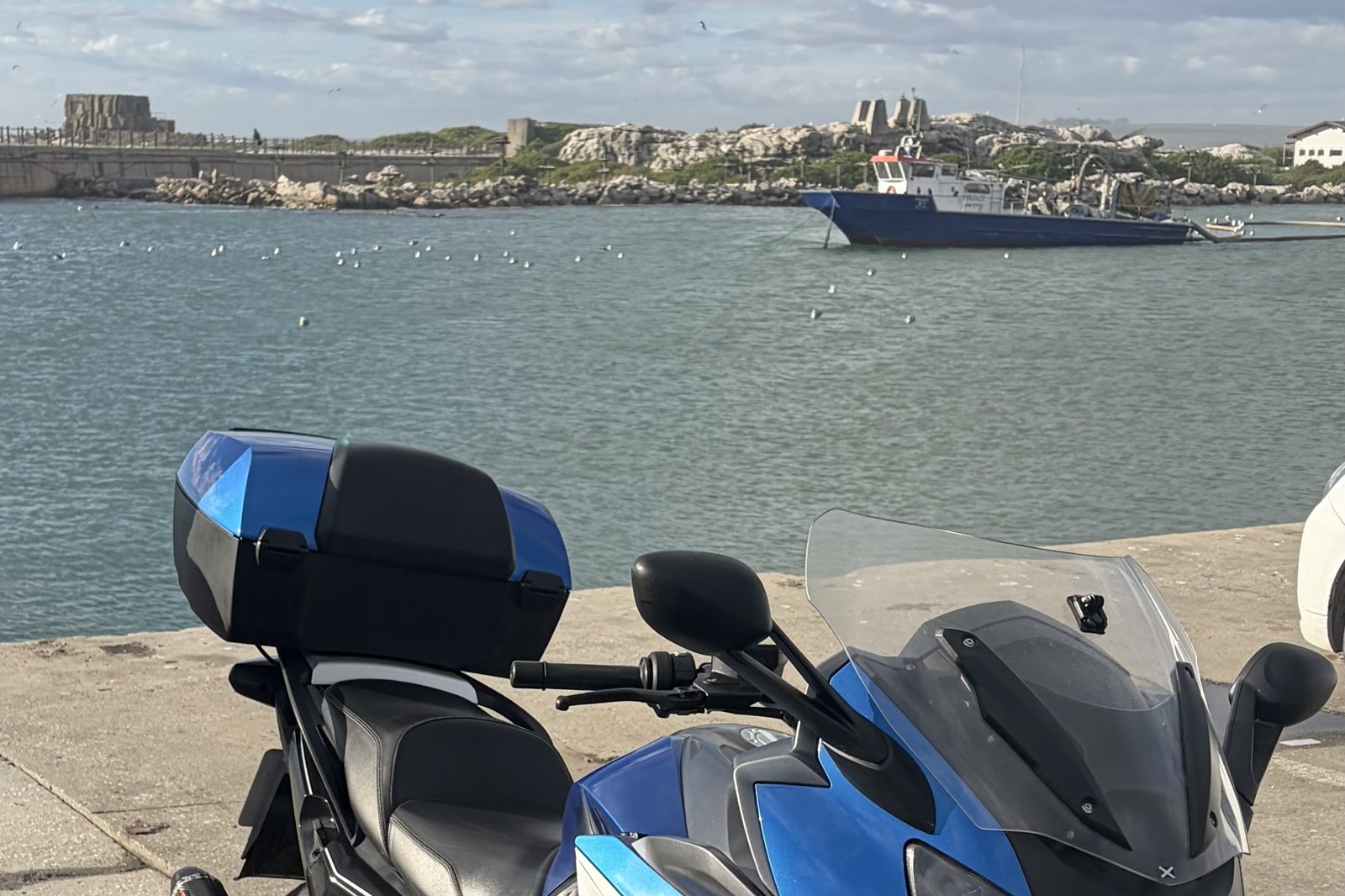 A BMW K1600 GT parked at a small fishing harbour on South Africa's West Coast, with a boat moored beyond.