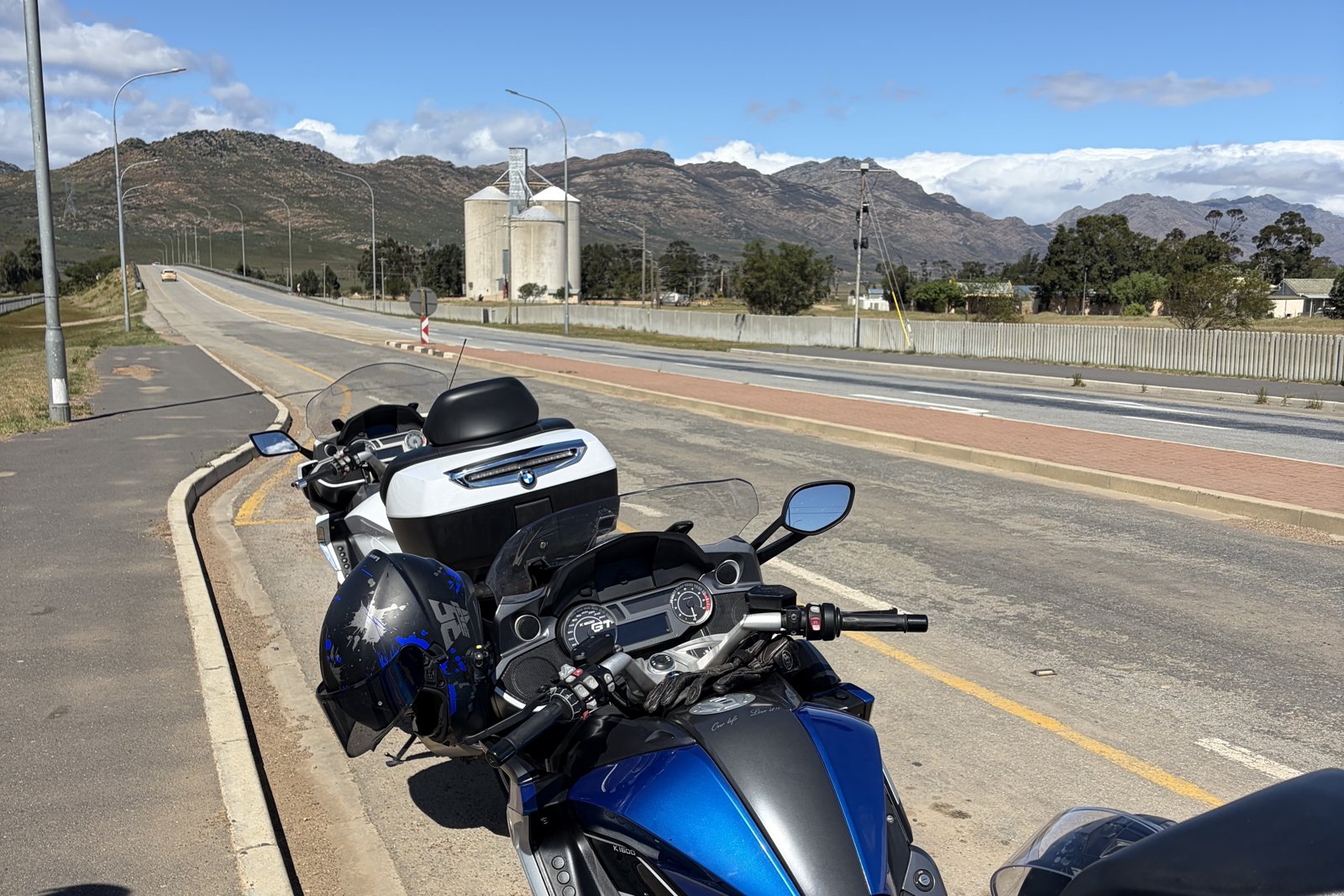 A BMW K1600 GT parked on a mountain pass road, with a grain silo and distant peaks behind.