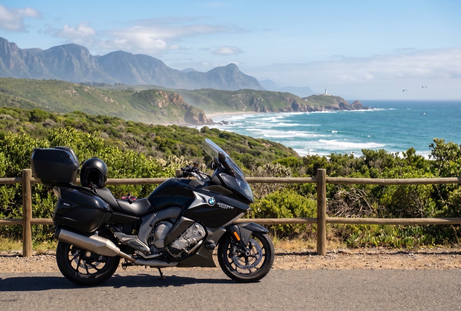 The same black BMW K1600 grand tourer parked above the Garden Route coastline, with green cliffs, breaking surf and a lighthouse beyond.